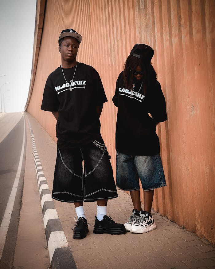 Two teens in streetwear pose stylishly by an orange wall in an urban setting.